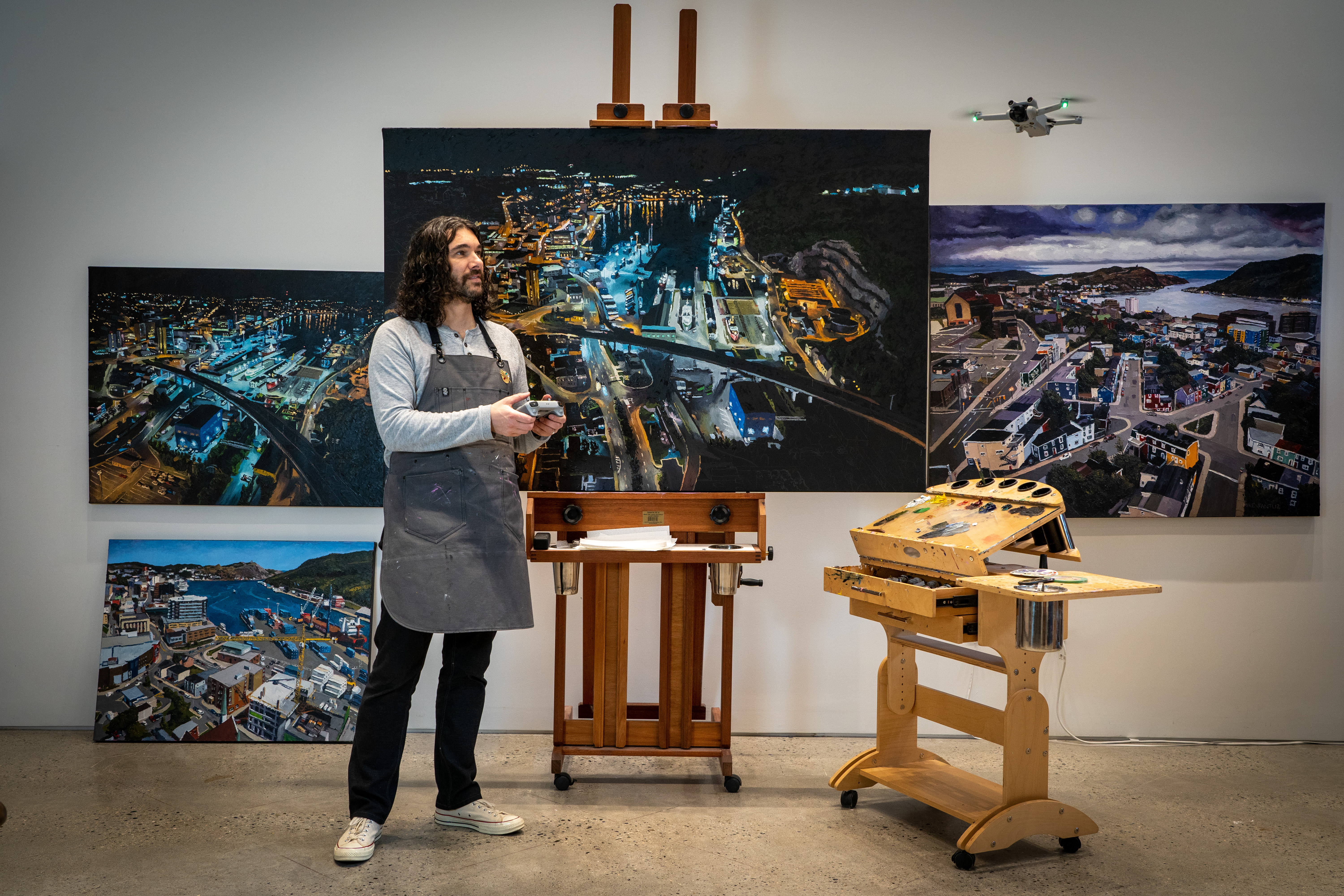 An artist flying a drone in front of various city-scape oil canvas paintings.